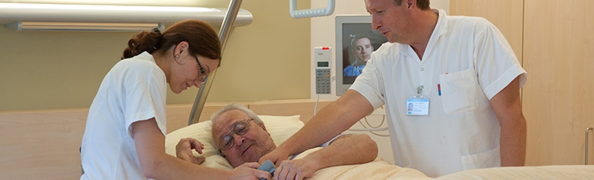 Einem im Bett liegenden Patienten wird die Steuerung zur Bedienung des Bettes von zwei Pflegepersonen erläutert