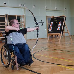 Ein Rollstuhlfahrer hat seinen Bogen gespannt und visiert das Ziel für den treffsicheren Schuss an. Das Training ist in der Sporthalle vom Rehabilitationszentrum.