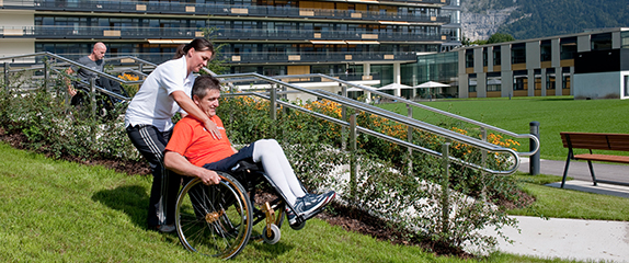 Therapeutin übt mit einem Patienten im Rollstuhl mit Unterstützung einen kleinen Grashang hinunter zufahren