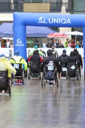 Im Bild sieht man eine Teilnehmergruppe von hinten durch den blauen Uniqa-Bogen fahren. An den Rollstühlen sind Startnummern fixiert. Alle tragen Regenschutz. Die Fahrbahn spiegelt sich vor Nässe.