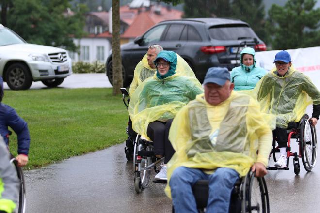 Drei Rollstuhlfahrerinnen und zwei Rollstuhlfahrer unterwegs. Dick eingepackt in warmen Jacken mit Kapuzen oder Käppis, darüber tragen sie den gelben Regenschutz. Starkregen und Kälte zieht ein.