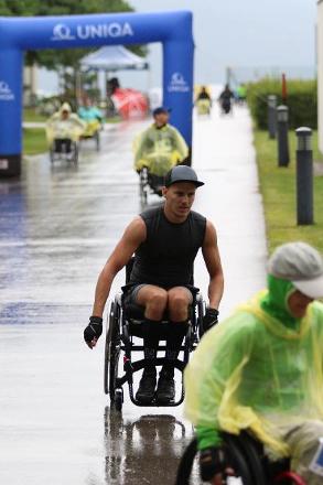Ein weiterer junger Rollstuhlfahrer ist auf der nassen Fahrbahnstrecke unterwegs. Ohne Regenschutz. Ein Käppi alleine schützt ihn vor dem Regen. In seinem Trägershirt und kurzer Hose blickt er konzentriert nach vorne.